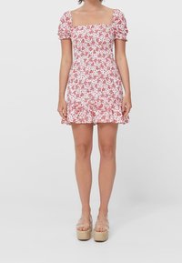 Blumendruckkleid in Weiß und Rot mit quadratischem Ausschnitt, kurzen Puffärmeln, gerafftem Taillenbund und ausgestelltem Saum. Getragen mit klaren Plattformsandalen.