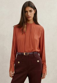 Bluza rjave oranžne barve s plisiranim dizajnom, odprtim izrezom v obliki ključavnice in dolgimi rokavi. Kombinirana s temno burgundijastimi hlačami z velikimi žepi.