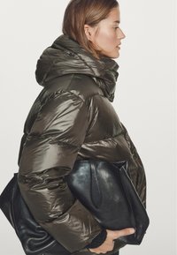 Kobieta w błyszczącej ciemnooliwkowej kurtce puchowej trzymająca dużą czarną skórzaną torbę typu tote, ukazana z profilu na jednolitym tle.