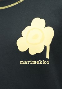 Γραφικό κίτρινο στυλιζαρισμένο λουλούδι με κοτσάνι και κεντρική λεπτομέρεια σε μαύρο ύφασμα, με το κείμενο "marimekko" κάτω από το λουλούδι.