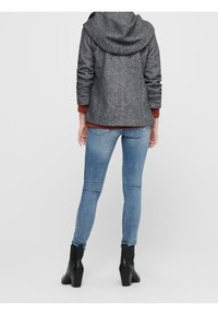 Giacca grigia con cappuccio e finitura texturizzata, indossata sopra una maglia color ruggine, abbinata a jeans skinny azzurri e stivaletti neri con tacco.