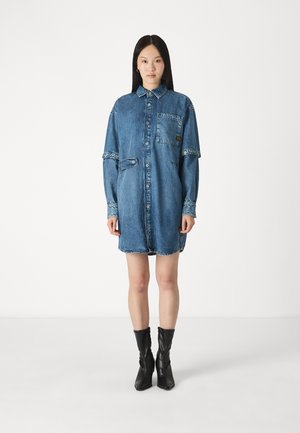 Jeanshemdkleid in Mittelblau mit Knopfverschluss, aufgenähtem Taschen und hochgerollten Ärmeln. Besitzt einen lockeren Schnitt und einen ausgefransten Saum.