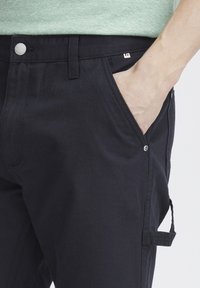 Los pantalones cargo negros tienen un bolsillo lateral, herrajes plateados y un cierre de botón en la cintura. Están hechos de una tela duradera con una textura suave.
