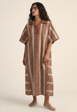 Mujer de pie descalza con vestido marrón holgado con franjas blancas cosidas verticales y horizontales y cuello tipo polo.