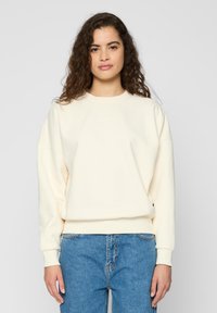 Krémová mikina s okrúhlym výstrihom, spustenými ramenami a rebrovanými manžetami. Obsahuje decentné vyšívané logo na hrudi. V kombinácii s modrými džínsmi.