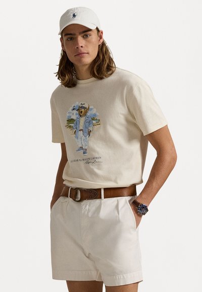 Beige bomulds t-shirt med et bjørne grafik, parret med hvide shorts. Oplægget har et brunt læderbælte og et ur. En hvid kasket fuldender looket.