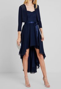 Marinblå klänning med åtsittande överdel, hög-låg chiffongsläpp och matchande kort jacka prydd med rhinestones. Silvriga strappar med klack och clutch.