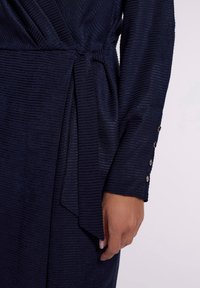 Robe portefeuille bleu marine à rayures texturées, avec une taille nouée, des manches longues et des accents de poignets ornés de boutons dorés.