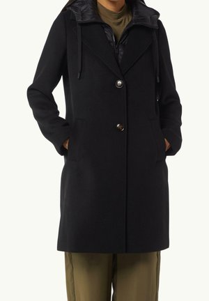 Manteau noir mi-long avec deux boutons, capuche avec cordons, poches latérales, porté sur un pantalon vert olive et un haut.