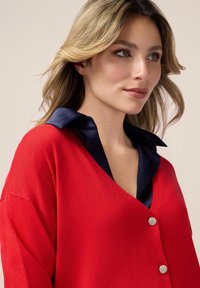 Gilet en tricot rouge avec un décolleté en V profond et deux boutons en argent. Doté d'un col en satin bleu marine. Texture douce, coupe décontractée.