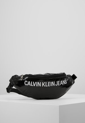 Kiiltävä musta Calvin Klein Jeans -vyölaukku, jonka etuosassa on suuri valkoinen logo, vetoketjukiinnitys ja säädettävä musta hihna, esillä valkoisella pinnalla.