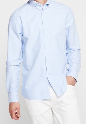 Camisa de manga larga azul claro con botones, usada sin meter en los pantalones por una persona con pantalones blancos, con una mano en el bolsillo, sobre un fondo liso.