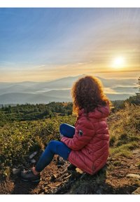 Osoba siedząca na skalistym terenie, ubrana w różową puchową kurtkę, niebieskie spodnie i ciemne buty, z malowniczym wschodem słońca nad górami w tle.