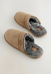 Pantofole beige testurizzate con una morbida fodera in pelo sintetico grigio, design leggero e un'etichetta distintiva sulla parte superiore. Utilizzo interno.