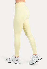 Högmidjade gula leggings i mjukt, elastiskt tyg med kontrastsömmar, kombinerade med vita sportskor.