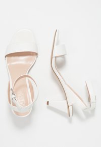Bílé sandály na vysokém podpatku se hladkým povrchem, s širokými pásky a špičatým blokovým podpatkem. Součástí je nastavitelný pás na kotníku.