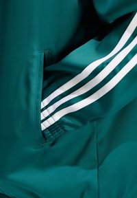 Teal urheilu takki, jossa on sileä rakenne, sivutaskut ja kolme valkoista raitaa hihassa korostamassa yksityiskohtia.