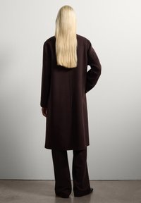 Lungo cappotto marrone realizzato in un tessuto texture, con taglio dritto, apertura posteriore e abbinato a pantaloni marroni coordinati.