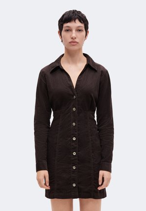 Femme aux cheveux courts et foncés portant une robe noire ajustée à manches longues avec col et boutons, sur un fond clair uni.