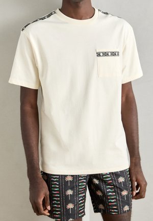 Roomkleurig t-shirt met korte mouwen en een borstzak, voorzien van een patroonaccent langs de schouders, gecombineerd met zwarte shorts versierd met kleurrijke palmboomontwerpen.