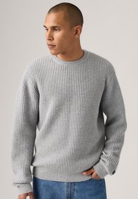 Grå strikket genser med ribbestrikkede ermer og hem, med et teksturert vaffelmønster. Stylet med blå denimbukser.