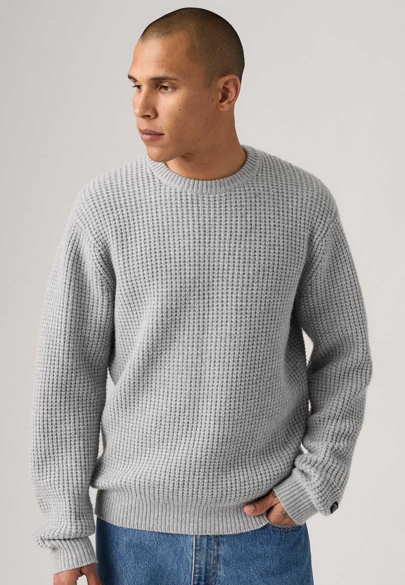 Grå strikket genser med ribbestrikkede ermer og hem, med et teksturert vaffelmønster. Stylet med blå denimbukser.