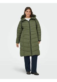 Zaļš pūkains mētelis ar kapuci, ar priekšējo rāvējslēdzēju, quilted dizainu un garajām piedurknēm, kombinēts ar tumšām džinsām un melnām zābakiem.