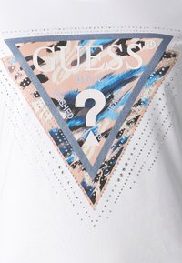 Camiseta blanca con un gran logo triangular que presenta elementos gráficos en azul, rosa y negro, adornado con strass a lo largo de los bordes.