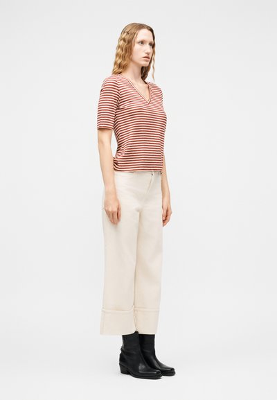 Γυναίκα με σγουρά ξανθά μαλλιά, ντυμένη με κόκκινη και λευκή ριγέ μπλούζα με V λαιμόκοψη, φαρδιά κρεμ παντελόνια με μανσέτες και μαύρα μποτάκια αστραγάλου, όρθια μπροστά σε λευκό φόντο.