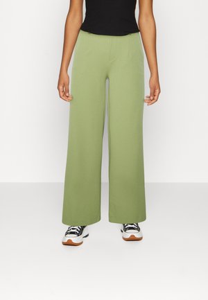 Person wearing olive green wide-leg pants, black sleeveless top, and black-and-white sneakers, standing against a white background.