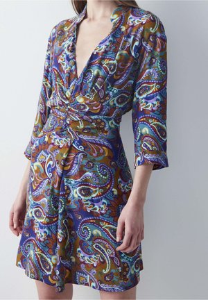 Frau trägt ein knielanges Wickelkleid mit einem bunten Paisley-Muster in Blau-, Lila-, Grün- und Brauntönen sowie Dreiviertelärmeln.