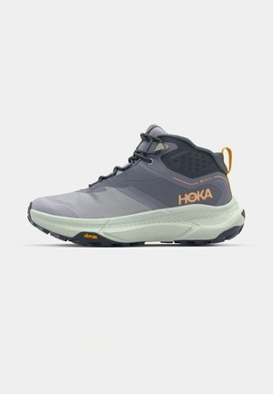 Scarpa da trekking HOKA grigia con tomaia testurizzata, dettagli sintetici e suola intermedia di colore verde chiaro. Presenta una suola Vibram per garantire aderenza.