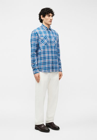 Άνδρας που στέκεται, φορώντας μπλε πουκάμισο με καρό μοτίβο και μακριά μανίκια, λευκό παντελόνι και σκούρα καφέ παπούτσια, μπροστά σε απλό ανοιχτόχρωμο φόντο.