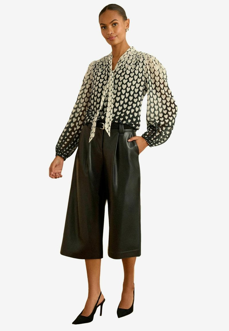 Blusa con motivo a cuori bianco e nero, con nodo al collo e maniche a sbuffo, abbinata a culotte in ecopelle nera a vita alta e tacchi neri.