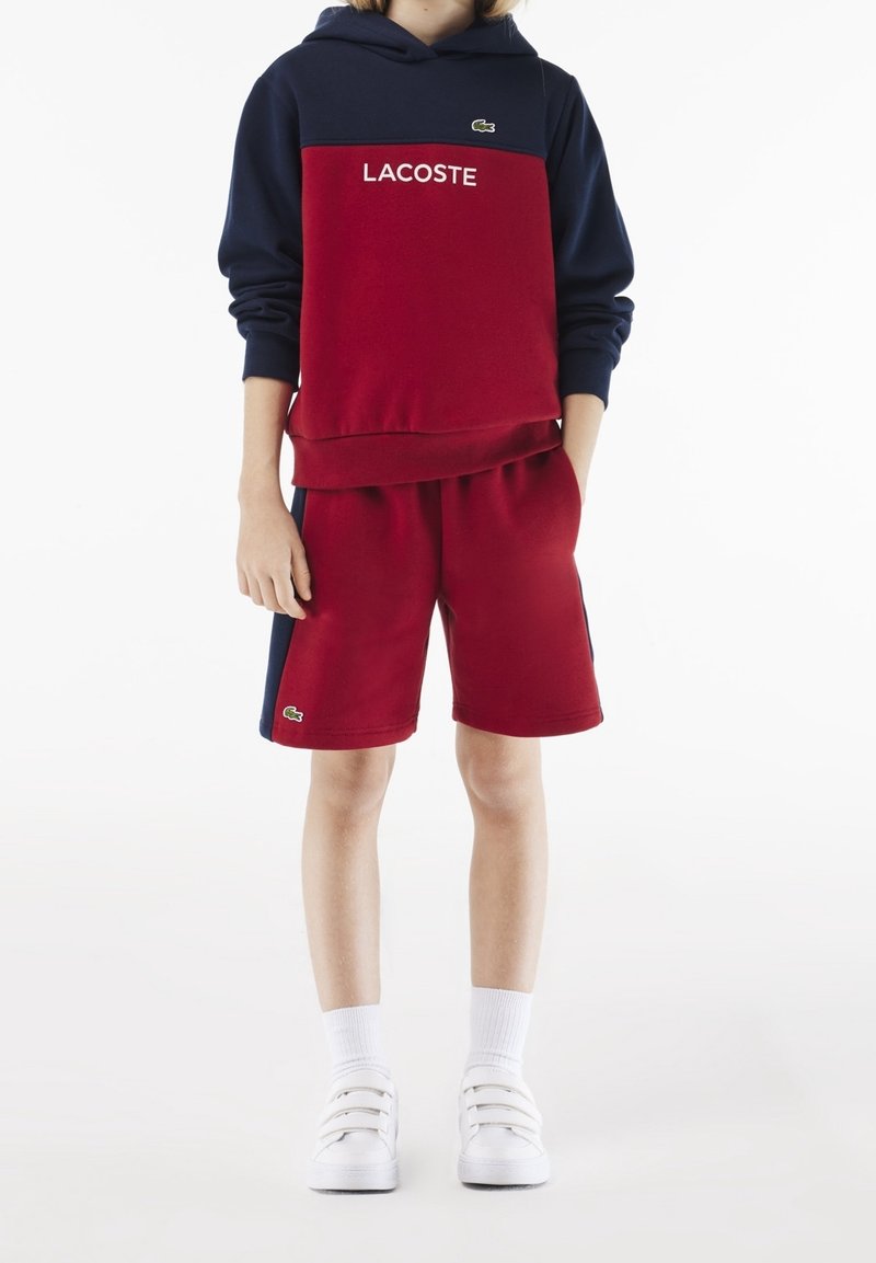 Felpa con cappuccio a blocchi di colore rosso e blu navy, con il logo "LACOSTE". Abbinata a pantaloncini rossi e sneaker bianche slip-on.