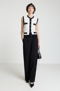 Weißes ärmelloses Strickweste mit schwarzen Akzenten und goldenen Knöpfen, kombiniert mit schwarzen Hosen mit weitem Bein und schwarzen spitzen Absatzschuhen.