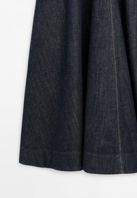 Tissu en denim bleu foncé avec des coutures visibles verticales et horizontales et une couture qui descend le long du côté droit sur un fond blanc.