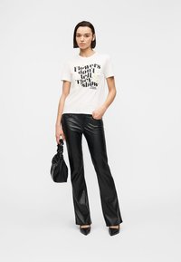 Γυναίκα που φοράει λευκό T-shirt με το κείμενο «Τα λουλούδια δεν λένε, δείχνουν» και μαύρο δερμάτινο παντελόνι, κρατώντας μια μαύρη τσάντα, στέκεται σε λευκό φόντο.