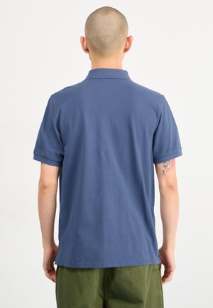 Mand med barberet hoved iført blå polo med korte ærmer og grønne bukser, vist bagfra mod en hvid baggrund.