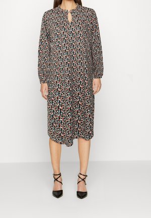 Kvinde iført en knælængde, kjole med lange ærmer og et flerfarvet abstrakt mønster samt nøglehulssnit ved halsen, kombineret med sorte hæle med ankelrem.