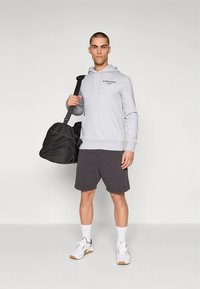 Sweatshirt cinzento com capuz e um bolso frontal, detalhe com logótipo, mochila de ginásio preta, calções escuros, meias brancas e ténis desportivos com detalhes em castanho-claro.