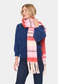 Bufanda de lana con patrón de cuadros en rojo, rosa y blanco. Textura suave con bordes desflecados. Llevada con un suéter azul de textura suave y jeans oscuros.