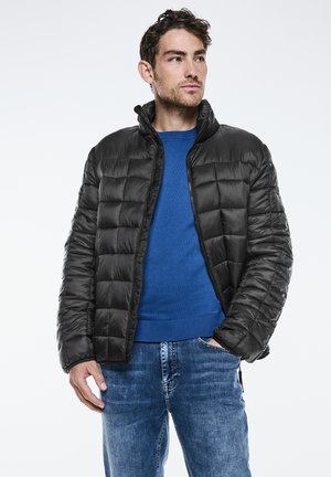 Chaqueta acolchada negra con cuello alto, diseño acolchado y cremallera frontal. Llevada sobre un suéter azul y combinada con unos vaqueros azules.