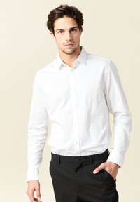 Camisa blanca de manga larga con botones, hecha de algodón, que presenta un cuello y puños estándar, combinada con pantalones negros texturizados.