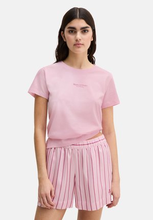 Frau mit langem, dunklem Haar, die ein hellrosa Kurzarm-T-Shirt und hellrosa gestreifte Shorts trägt und vor einem schlichten Hintergrund steht.