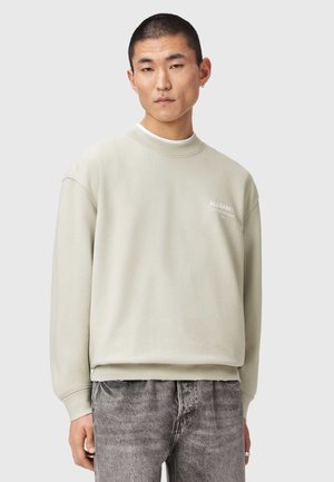 Man draagt een licht beige AllSaints-trui en grijs gewassen spijkerbroek, staand tegen een effen lichtgrijze achtergrond.