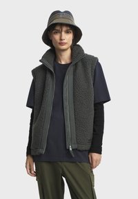 Gilet en polaire gris avec un col haut et une fermeture éclair à l'avant, porté par-dessus une chemise marine et un pantalon vert. Accompagné d'un chapeau seau à motif.