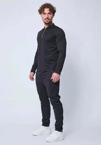 Haut noir à manches longues avec un col zippé, associé à un pantalon de jogging noir fuselé et des baskets blanches. Tissu lisse et design ajusté.