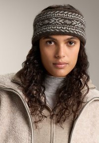 Un bandeau en tricot marron et gris, présentant un motif texturé. Porté avec une veste en polaire zippée beige clair sur un col roulé à côtes.