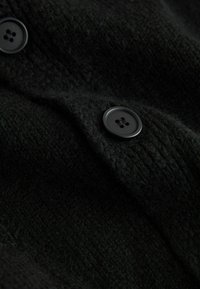 Zwart gebreide stof met een gestructureerd oppervlak en twee zwarte knoopsluitingen. Het materiaal lijkt zacht en dik.